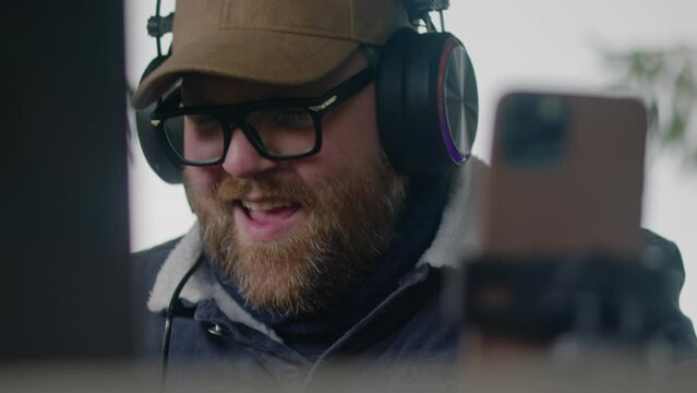 Close up of Caucasian musician in headphones playing MIDI keyboard, creates music and moves rhythmically sitting in gazebo in forest. Adult man uses laptop and phone on tripod to record song outdoors.