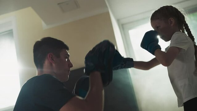 Instructor and child fight at sparring in gym. Careful trainer with glove pads trains girl in sports club slow motion. Physical activity for kids