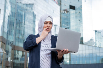 Fototapeta premium Thoughtful business woman in hijab and suit is holding a laptop outdoors.