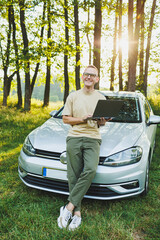 A cheerful man in glasses sits on the hood of his car in nature and works on a laptop. Freelance...