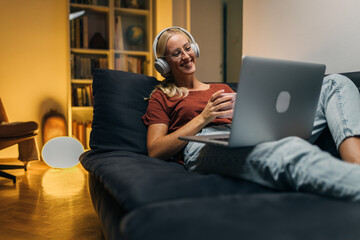 Smiling woman with headset sitting on the sofa and looking at laptop.