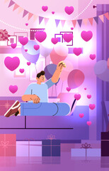 guy in love sitting on sofa in living room with gift boxes and pink air balloons in heart shape happy valentines day celebration concept