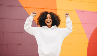 Teenage girl wearing a white hoodie with no print or logo standing in front of colorful wall, raising hands in excitement, apparel mockup photo, African-American child