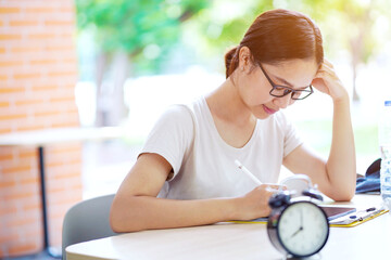 Asian young female student in casual cloth doing homework with alarm clock on table at her school. concept of time management - Powered by Adobe