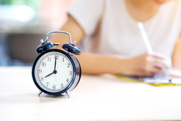 Selective focus of alarm clock on table near young female student in casual cloth doing homework at her school. concept of time management. - obrazy, fototapety, plakaty