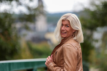 An older blond woman wearing a brown leather jacket looks over her shoulder and laughs at the camera - obrazy, fototapety, plakaty