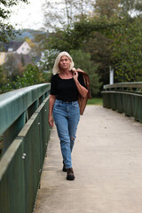 An older, blonde woman in a leather jacket and a black shirt strolls across a bridge and looks into the camera.