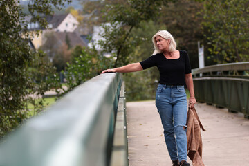 A woman in her mid-forties wearing a black shirt is holding on to a bridge railing with her arm...