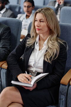 Vertical image of elegant businesswoman making notes in her notepad while visiting business conference