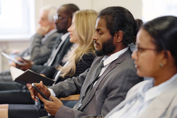 Young businessman in suit using mobile phone while sitting at conference with other people