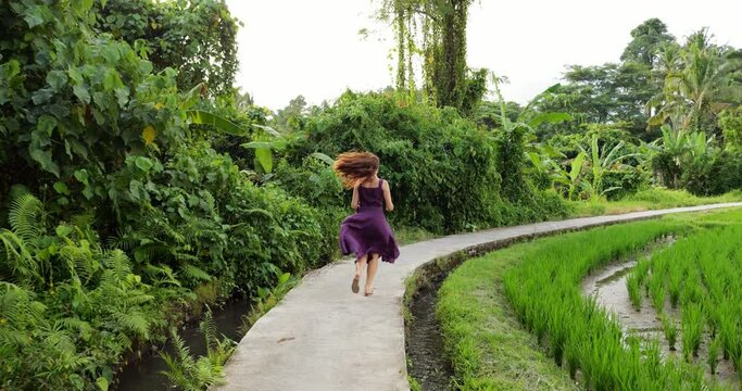 Happy woman running at bending footpath in Ubud backsides, lush green bushes on left and rice fields at right. Joyful tourist wearing dress with long skirt, it flying when she jog