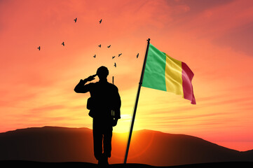 Silhouette of a soldier with the Mali flag stands against the background of a sunset or sunrise....