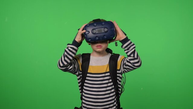 A little excited boy uses a virtual reality headset in a studio with a green background. The boy wears virtual reality glasses and turns his head around.