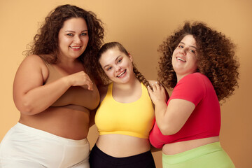 Elegant, positive, smiling young women, friends standing in sportswear over beige studio background. Oversize models. Concept of sport, body-positivity, weight loss, body and health care