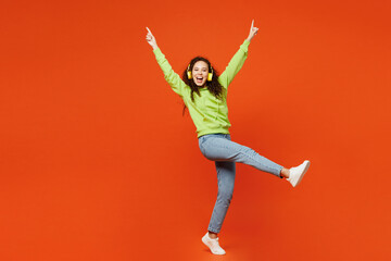 Full body happy young woman of African American ethnicity she wear green hoody casual clothes listen to music in headphones raise up hands isolated on plain red orange background. Lifestyle concept.