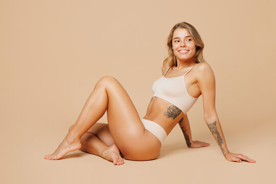 Full body side view happy fun young nice lady woman with slim body perfect skin wear nude top bra lingerie sitting looking aside isolated on plain pastel beige background. Lifestyle diet fit concept.