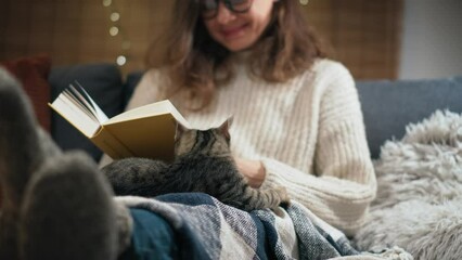 A young Caucasian woman reading a book sitting on the couch with her cute grey cat at home on a winter evening - Powered by Adobe