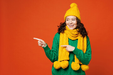 Young happy woman she wear green knitted sweater yellow hat scarf pointing index finger aside indicate on workspace area copy space isolated on plain orange red background studio. Lifestyle concept.