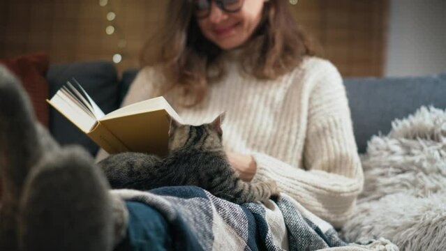 A young Caucasian woman reading a book sitting on the couch with her cute grey cat at home on a winter evening
