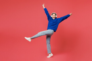 Full body cool young woman of Asian ethnicity she wear blue sweater casual clothes listen to music in headphones raise up leg spread hands isolated on plain pastel pink background. Lifestyle concept.