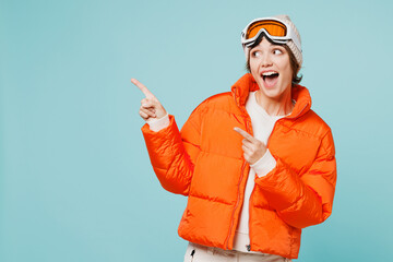 Skier young woman wearing warm padded windbreaker jacket hat ski goggles mask point index finger aside on area travel rest spend weekend winter season in mountains isolated on plain blue background. © ViDi Studio