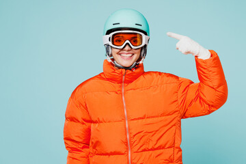 Young skier woman wear warm padded windbreaker jacket ski goggles mask point index finger on helmet look camera travel rest spend weekend winter season in mountains isolated on plain blue background.