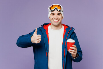 Skier man wear warm blue windbreaker jacket ski goggles mask hat hold takeaway cup hot coffee to go show thumb up spend extreme weekend winter season in mountains isolated on plain purple background.