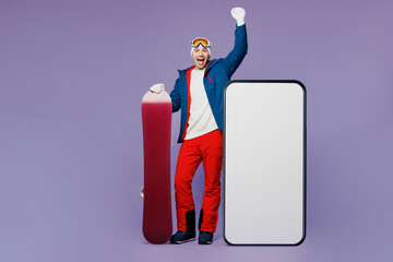 Full body winner man he wear blue jacket ski goggles mask hat hold snowboard big blank screen of mobile cell phone spend extreme weekend winter season in mountains isolated on plain purple background