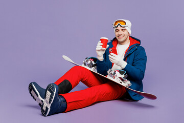 Full body fun man wear blue jacket ski goggles mask hat sitting hold snowboard drink coffee use...