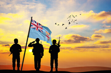 Silhouettes of soldiers with the Fiji flag stand against the background of a sunset or sunrise....
