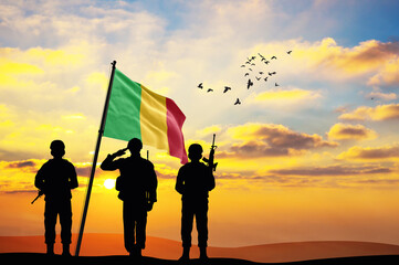 Silhouettes of soldiers with the Mali flag stand against the background of a sunset or sunrise....