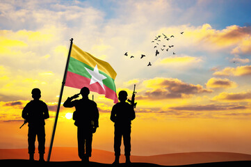 Silhouettes of soldiers with the Myanmar flag stand against the background of a sunset or sunrise....
