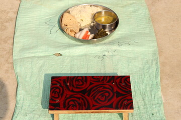 A steel plate thali with Normal Indian Staple food including radish, carrot, rice, chapati, daal, gajak, siting bench on ground