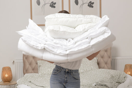 Woman holding pile of soft white folded duvet and pillows at home in her bedroom, cozy domestic lifestyle, housewife cleaning, tidying up bedroom, housework chores concept.