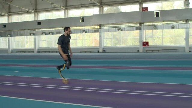 Slowmo of determined Caucasian sportsman with prosthetic running blade doing professional long jump on sandy pit at indoor stadium with track