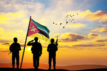 Silhouettes of soldiers with the Mozambique flag stand against the background of a sunset or...