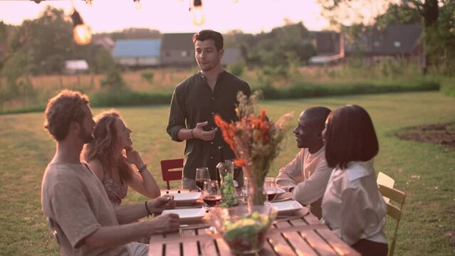 Man Telling A Story To His Guests Sitting At The Outdoor Garden Picnic Dinner Table At Sunset