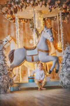Holiday. New Year. Christmas. Carousel. A boy in a white shirt and beige trousers sits near gifts and snow-covered Christmas trees. New Year's interior. The kid is standing by the carousel. A horse. S