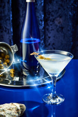 Glass filled with martini, garnished with two green olives small, silver tray with olives and bottle on it against blue background. Concept of night life, party time, celebration, holidays, New Year.