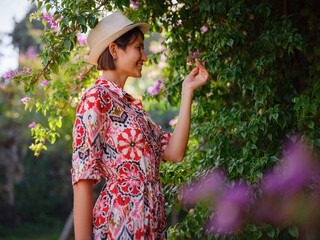 young asian woman stands in sunny green park, admiring blooming bougainvillea bush. The flower's...