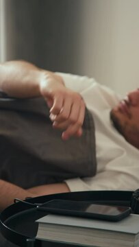 Vertical side stab of Caucasian bearded man waking up to alarm going off on charging phone and getting up from bed at home