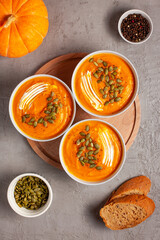 Three plates of pumpkin soup with seeds, sour cream, spices and bread on a gray background, shot from above. The concept of healthy vegetable soups. Concept of healthy eating or vegetarian food