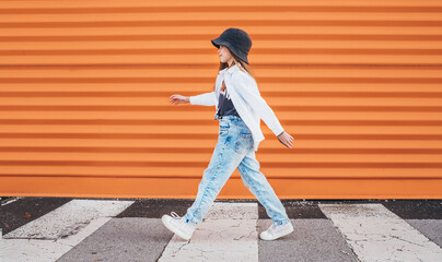 Little fashion-dressed girl funny walking crossing a pedestrian zebra crosswalk on the orange wall...