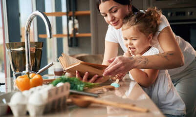 Holding book with recipe. Mother with her daughter are preparing food on the kitchen