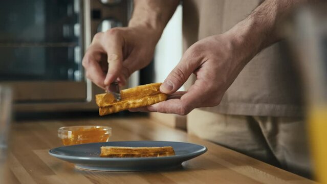 Side stab shot of male hands spreading apricot jam on toasted slice of bread standing in modern kitchen and preparing breakfast