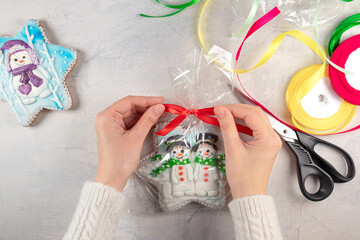 female hands in a white sweater tie a red bow on a package of gingerbread cookies with snowmen on the background of a light table, multi-colored ribbons and scissors. concept for self-made Christmas