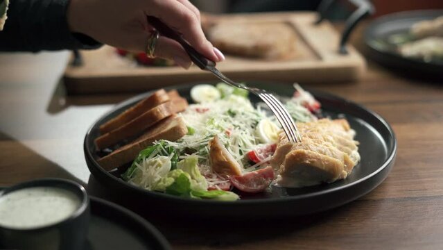Close-up of woman eating Caesar salad in the restaurant. Grilled chicken breast sliced, green salad leaves, croutons and Parmesan on plate with menu