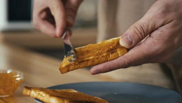 Closeup shot of unrecognizable man spreading orange marmalade on slice of toasted bread using knife while preparing breakfast in kitchen