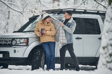 Man is covering woman with warm blanket. Beautiful couple standing near the car in the winter forest