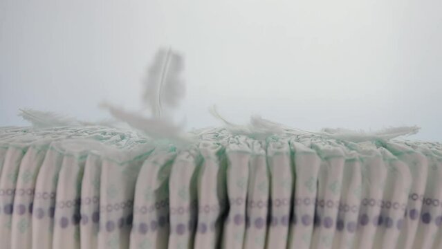 Feathers fall on a stack of baby diapers on a white background. The concept of softness and comfort, thorn-allergenic diapers made of cotton. Copy space for text. Slow motion